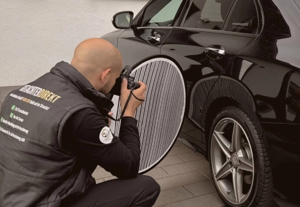 KFZ-Sachverständiger fotografiert Fahrzeugschaden an schwarzem PKW mit Dellenreflektor